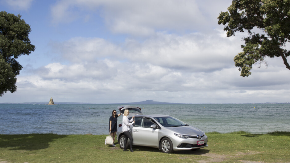 Two women standing outside GO Rentals vehicle parked on grass with Rangitoto Island in background.