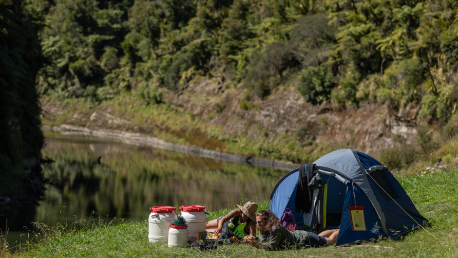 Paddlers relax at a camp overlooking the river with tent and storage drums