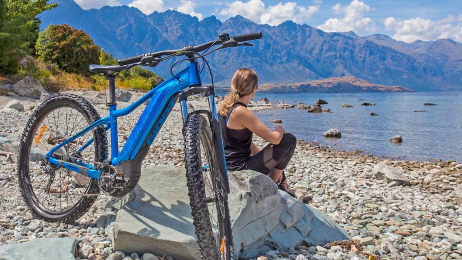 The Frankton track is easy and navigatable via following the edge of the lake.  The Remarkables stands tall in the distance. A customer enjoys the waves with a blue Ebike fills the foreground.