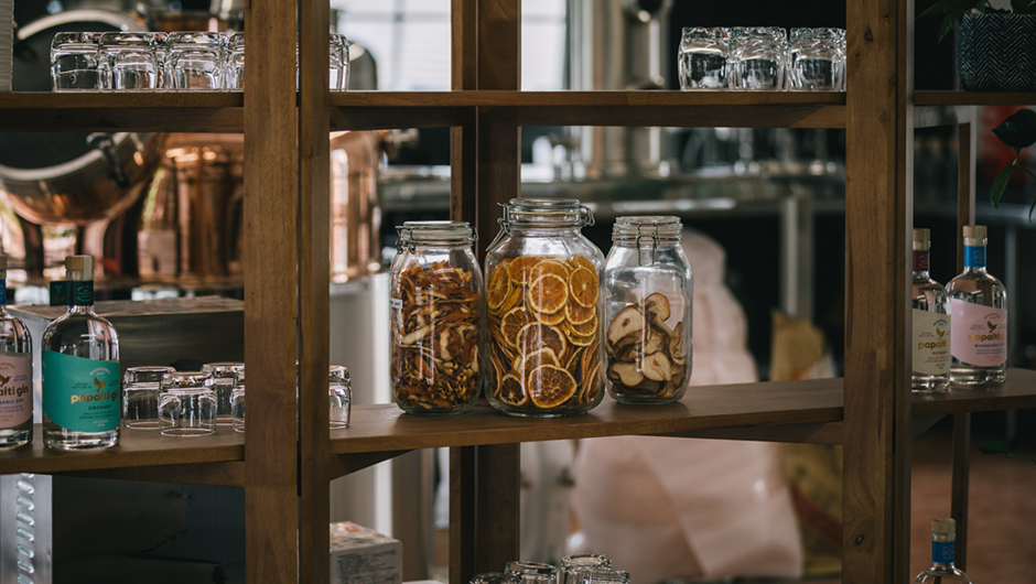 Jars of dehydrated botanicals sitting on a shelf in front of big copper still at Papaiti Gin Distillery.