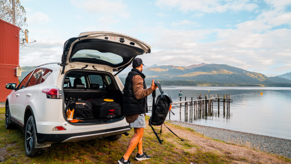 A male person grabbing his backpack from behind the GO Rentals vehicle boot with a lake and mountain landscape.