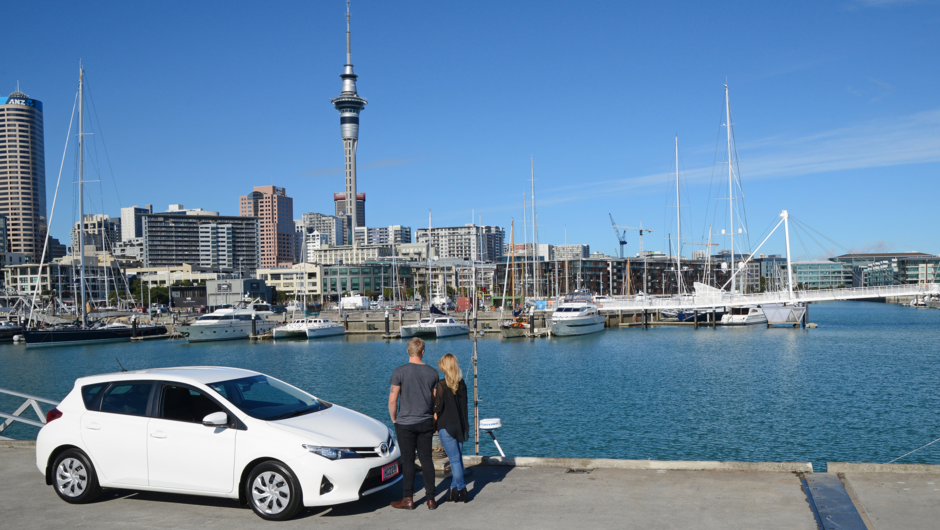 A couple standing in front of GO Rentals white vehicle by the wharf side with Auckland Tower and City background.