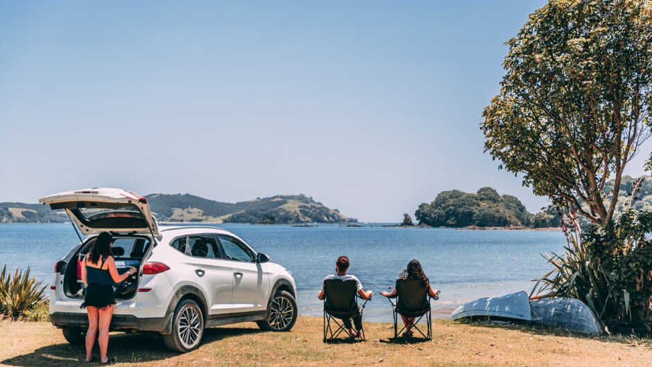 A person grabbing something from GO Rentals vehicle boot while two other people are sitting on camping chairs next to the vehicle admiring the beach and mountain landscape.