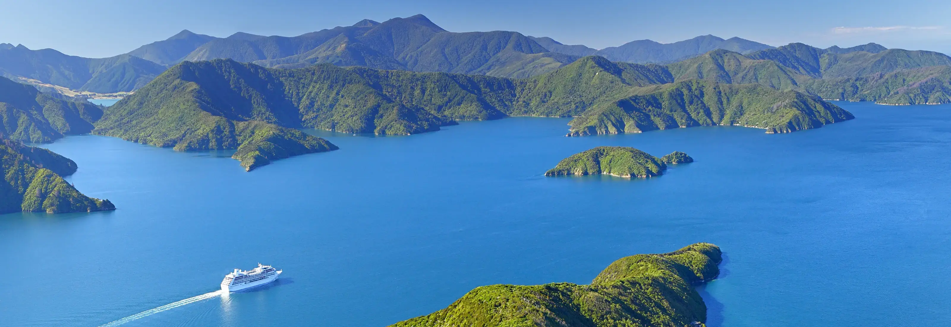 Spectacular Marlborough Sounds with pristine blue waters winding between lush green forested hills and secluded bays. A cruise ship navigates the intricate waterways of this stunning South Island fjord system in New Zealand.