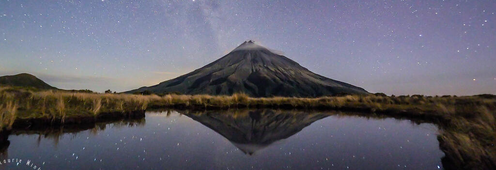 Looking across Pouakai Tarn towards Taranaki Maunga at night. Beautiful starscapes are reflected in the water.